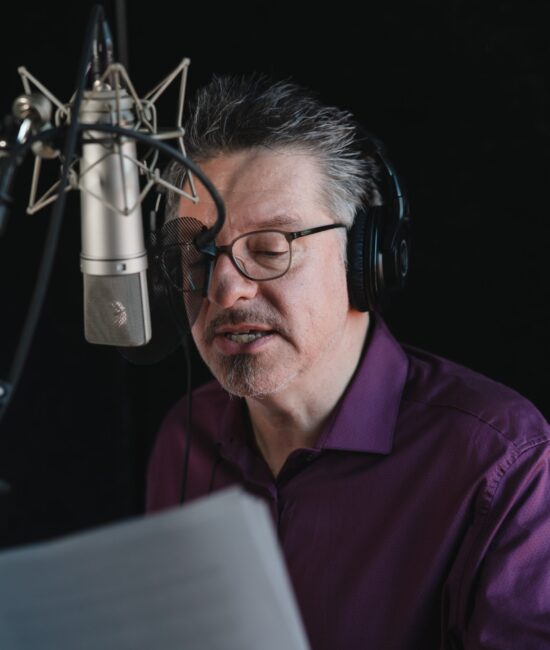 Homme portant des écouteurs devant un micro lisant un texte à enregistrer qu'il tent dans une main